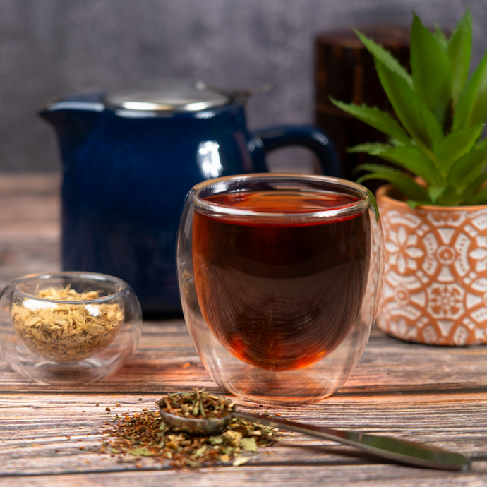 A double-walled glass filled with brewed Organic Fresh Start Tea on a wooden table, with a blue teapot, dried herbs, and a potted plant in the background.