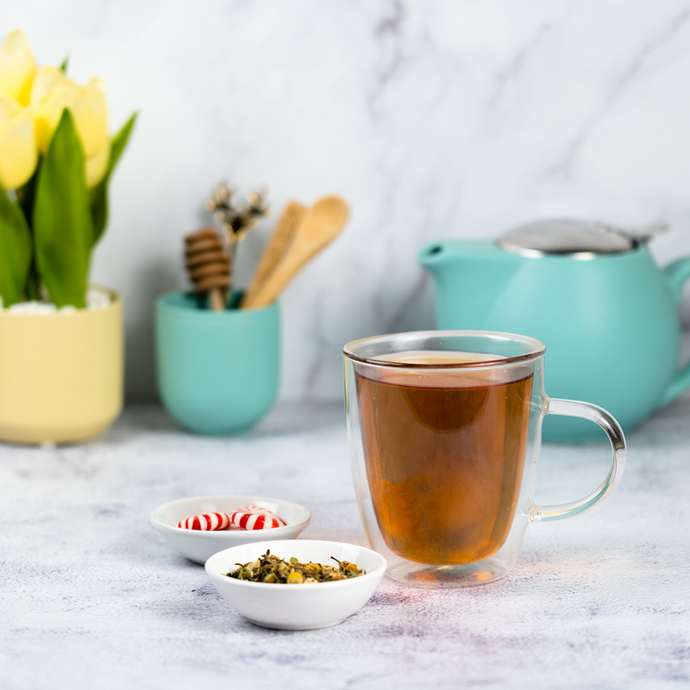 A clear glass mug of brewed Organic Fresh Start Tea beside small bowls of loose leaf tea and peppermint candies, with a turquoise teapot and potted plants in the background on a light surface.
