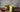 A refreshing iced matcha latte served in a mason jar with a glass straw, placed on a rustic wooden table. A small wooden bowl with matcha powder and a decorative plant in a geometric gold pot sit in the background. The text overlay reads 'Craft an Iced Ma