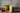 A refreshing iced matcha latte served in a mason jar with a glass straw, placed on a rustic wooden table. A small wooden bowl with matcha powder and a decorative plant in a geometric gold pot sit in the background. The text overlay reads 'Craft an Iced Ma