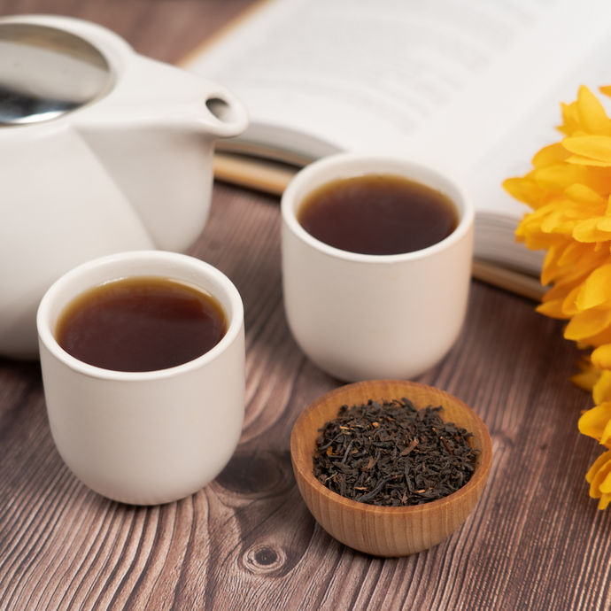 Two cups of Organic Earl Grey tea beside a white teapot, a wooden bowl of loose black tea, and an open book with yellow flowers on a wooden surface.