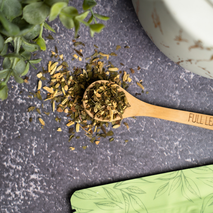 Organic Tummy Balance Tea loose blend on a bamboo spoon surrounded by fresh green herbs, featuring fennel, peppermint, and licorice root pieces on a dark stone surface beside a green Full Leaf Tea Co. package.