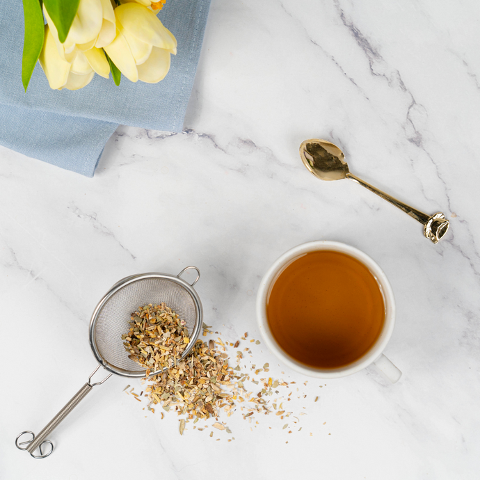 Cup of Organic Liver Cleanse tea with loose leaf herbs in a tea strainer on a white marble surface, accompanied by a gold spoon and yellow flowers.