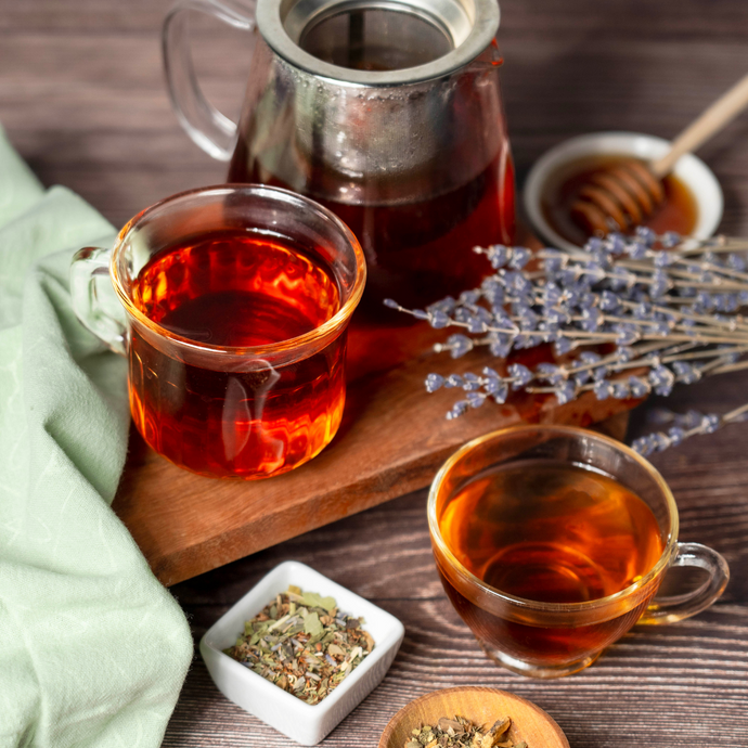 Organic Relieve Stress Tea brewed in a glass teapot and served in clear cups, surrounded by dried lavender, honey, and herbal tea leaves on a wooden surface.