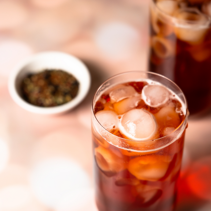 Glass of iced Organic Immunity Blend tea filled with ice cubes, with loose leaf tea in a small dish in the background on a soft pink surface.