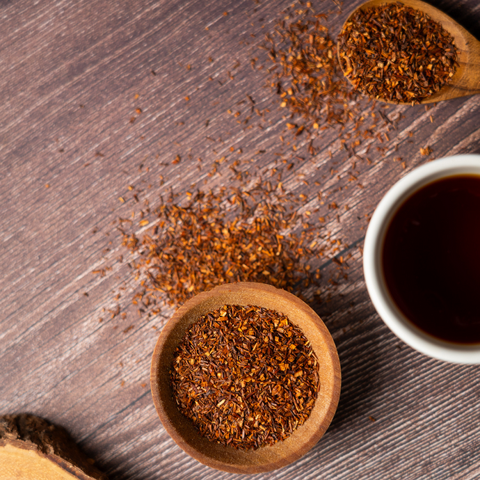 Organic Rooibos Tea leaves in a wooden bowl and spoon with some scattered on a rustic wooden surface, beside a cup of freshly brewed red rooibos tea.