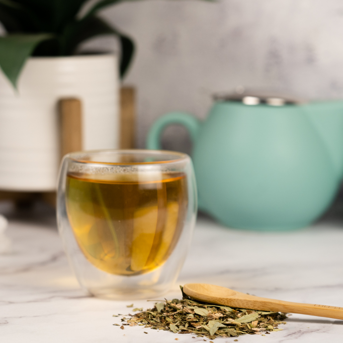 Glass of brewed Organic Healthy Colon Tea beside a wooden spoon resting on a small pile of loose herbal tea leaves, with a mint-green teapot and potted plant in the background.