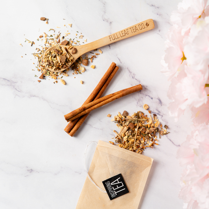Flat lay of loose leaf herbal tea with cinnamon sticks, wooden spoon engraved with ‘Full Leaf Tea Co.’, and a tea bag labeled ‘Inspiring Tea’ on a white marble surface with pink flowers along the edge.