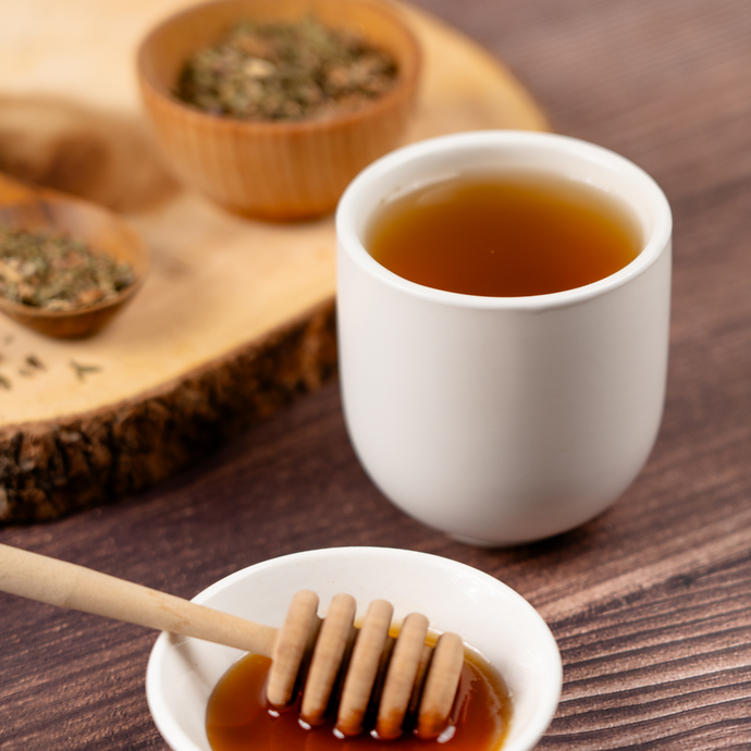 Cup of Organic Dandelion Tea on a wooden surface beside a small dish of honey with a honey dipper, and bowls of loose dried herbs in the background on a wood slab.