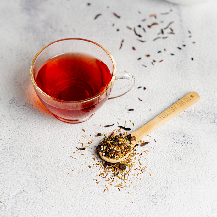 Cup of freshly brewed Organic Vita Me Tea with a deep red color beside a bamboo spoon of loose herbal tea blend on a light stone surface, with scattered tea leaves around.