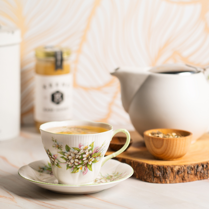 A floral teacup filled with Organic Kidney Cleanse tea sits on a marble surface beside a white teapot, a small wooden bowl of herbal tea blend, and a jar of honey in the background.