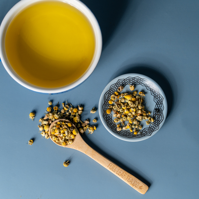 A cup of Organic Chamomile Tea beside a wooden spoon and small dish of dried chamomile flowers on a blue surface, showcasing a calm and minimal aesthetic.