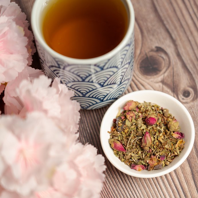 Cup of herbal tea with a patterned blue and white mug beside a bowl of loose leaf tea with rosebuds, surrounded by pink flowers on a wooden table.