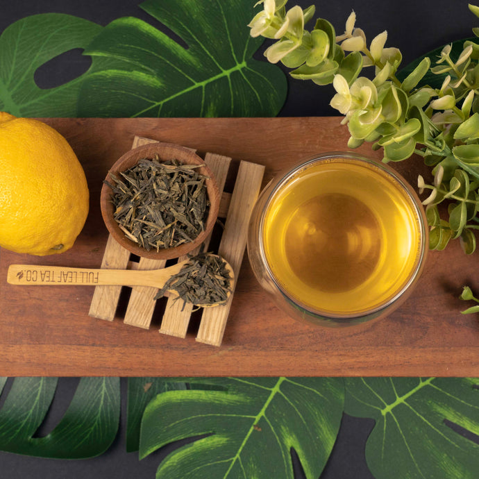 Tea-making setup with a cup of tea, dry tea leaves, a lemon, and a wooden spoon on a wooden board with green leaves in the background.