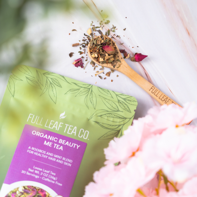 Full Leaf Tea Co. Organic Beauty Me Tea package with wooden spoon of loose leaf rooibos and mint blend, surrounded by pink flowers on a light marble background.