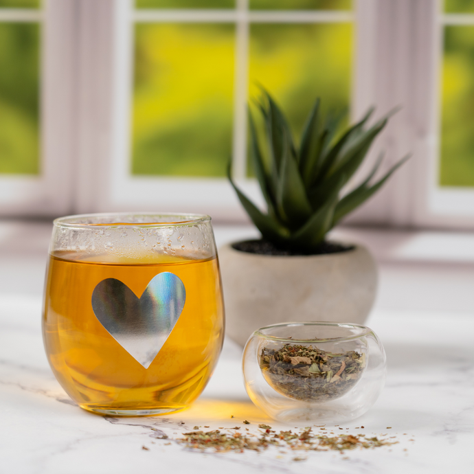 Glass cup of Organic Detox Tea with a heart design beside a smaller glass bowl of loose tea leaves, placed on a marble surface with a green plant and window in the background.