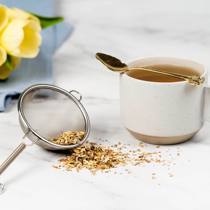 Cup of Organic Liver Cleanse Tea with a gold spoon, loose leaf herbs, and a metal tea strainer on a marble surface beside yellow flowers.