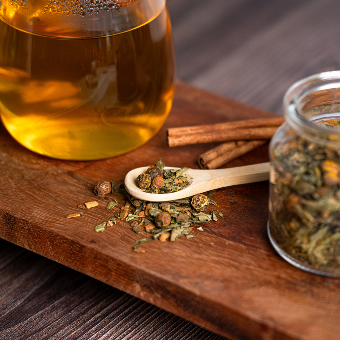 Close-up of Organic Healthy Heart Tea with loose leaf blend on a wooden spoon beside a glass mug of brewed tea, a jar of herbs, and cinnamon sticks on a wooden board.