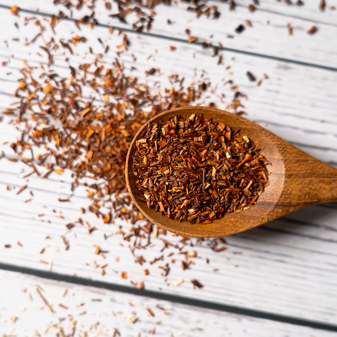 Close-up of loose Organic Rooibos Tea in a wooden spoon with scattered red-brown tea leaves on a light wooden surface.