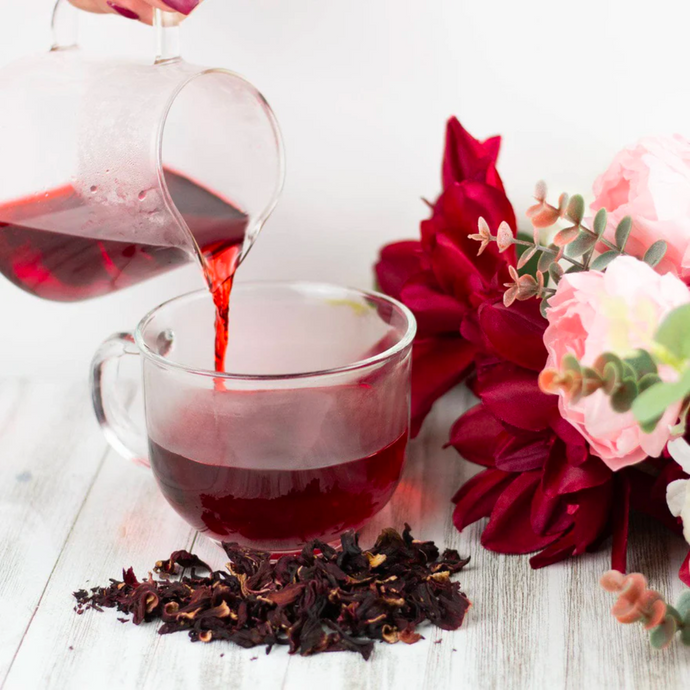 Bright red Organic Hibiscus Tea being poured from a glass pitcher into a clear teacup, with dried hibiscus petals on a light wooden surface and pink and burgundy flowers arranged beside it.