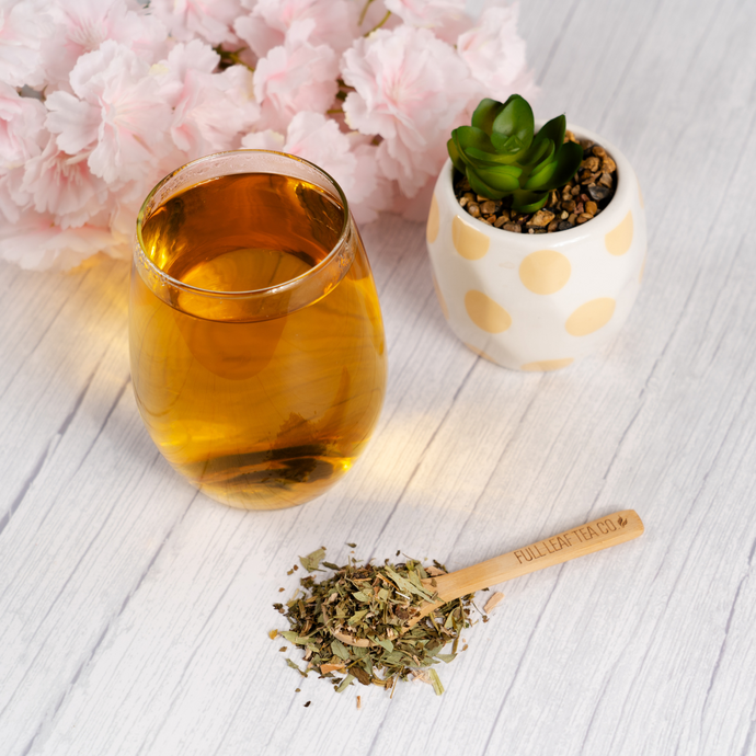 Glass of brewed Organic Healthy Colon Tea beside a bamboo spoon filled with loose tea leaves, pink flowers, and a small succulent on a light wood surface.