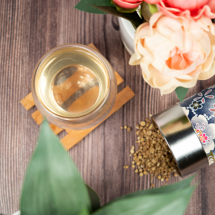 A glass cup of Organic Astragalus Tea on a wooden coaster surrounded by flowers and greenery, with a decorative tin spilling dried astragalus root pieces on a wooden surface.