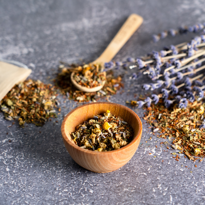 Loose leaf Organic Sleeping TranquiliTea blend with chamomile and lavender in a wooden bowl, with a bamboo spoon, tea bag, and dried lavender flowers on a gray stone surface.