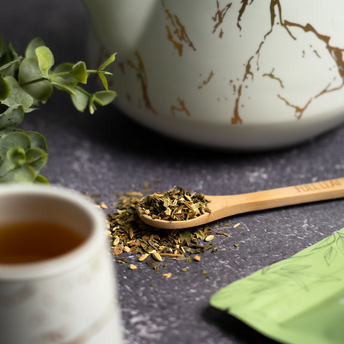 Organic Tummy Balance Tea loose blend on a bamboo spoon beside a cup of brewed tea and a white teapot with gold marbling, set on a dark stone surface with a green Full Leaf Tea Co. package.