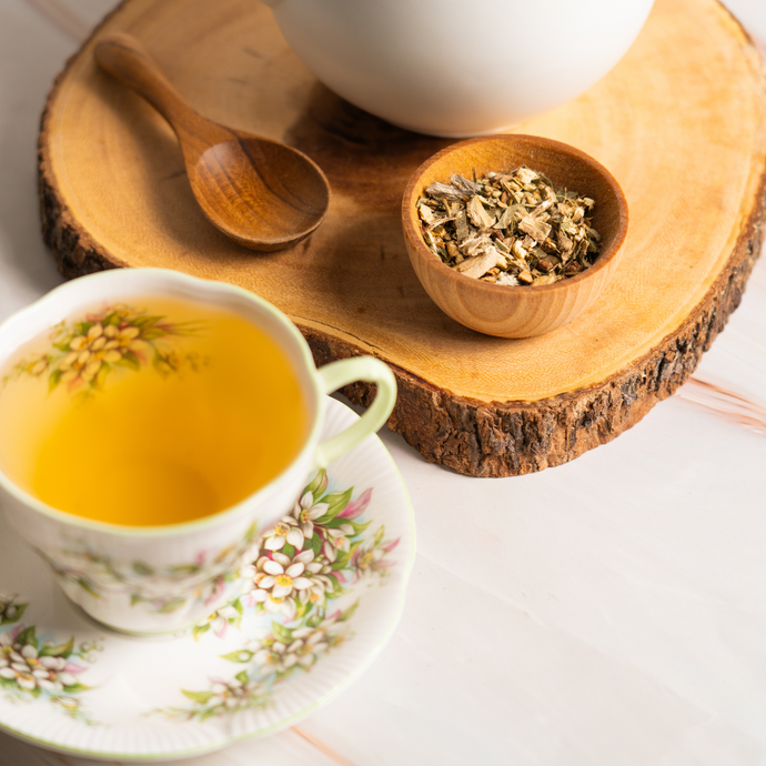 A floral teacup filled with bright golden Organic Kidney Cleanse tea beside a small wooden bowl of dried herbal blend and a wooden spoon on a natural wood slice tray.