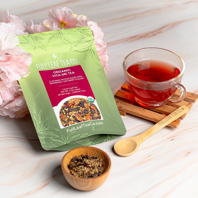 Full Leaf Tea Co. Organic Vita Me Tea package displayed beside a glass cup of vibrant red brewed tea, a bamboo spoon, and a small wooden bowl of loose tea leaves on a light wooden surface with pink flowers.