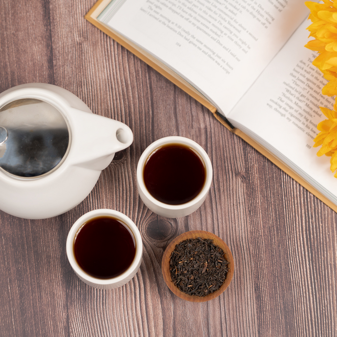 Top view of Organic Earl Grey tea served in white teacups beside a teapot, loose black tea leaves in a wooden bowl, and an open book with yellow flowers on a wooden table.