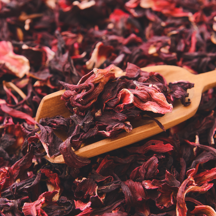 Close-up of dried hibiscus petals with deep red and magenta hues, spilling from a wooden scoop onto a pile of loose hibiscus tea