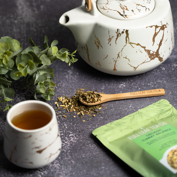 Cup of brewed Organic Tummy Balance Tea beside a white teapot with gold marbling, a bamboo spoon of loose tea blend, and a green Full Leaf Tea Co. package on a dark stone surface.