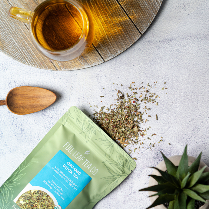 Flat lay of Full Leaf Tea Co. Organic Detox Tea with loose tea leaves spilled beside a wooden spoon, a glass mug of brewed tea, and a small plant on a light textured surface.