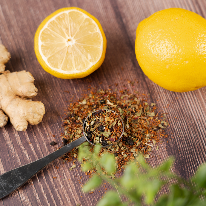 Organic Immunity Blend loose leaf tea with a metal teaspoon surrounded by fresh ginger, lemon, and green herbs on a wooden surface.
