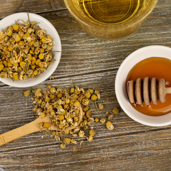 A cup of Organic Chamomile Tea with dried chamomile flowers on a wooden spoon, a small bowl of honey with a honey dipper, and a rustic wooden surface beneath.