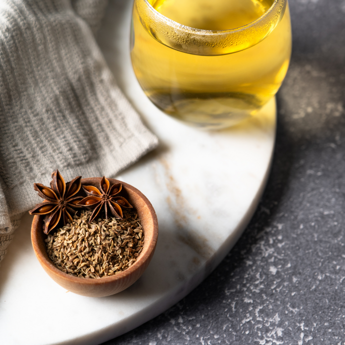 A cup of Organic Anise Seed Tea on a marble board beside a small wooden bowl filled with dried anise seeds and star anise pods, with a soft gray cloth in the background.