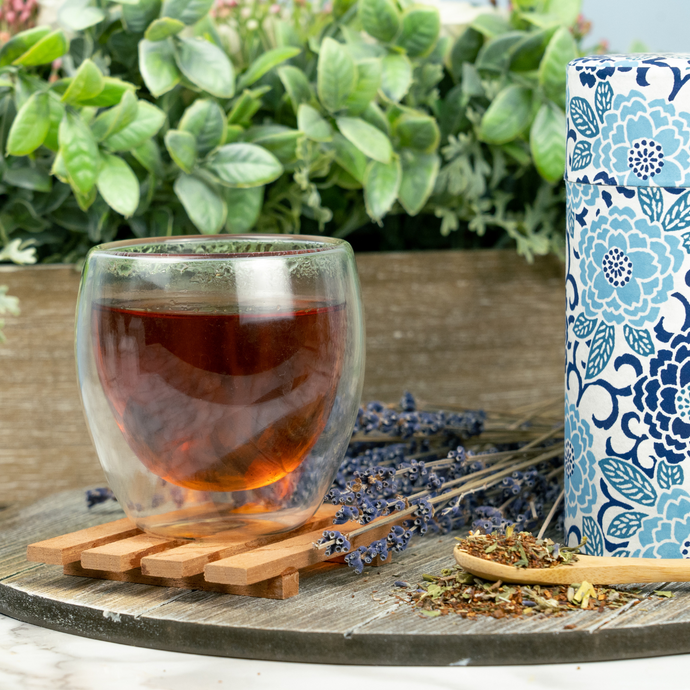 Glass cup of Organic Relieve Stress Tea with deep amber color, dried herbs and lavender on a wooden tray, and blue floral tea tin in the background.