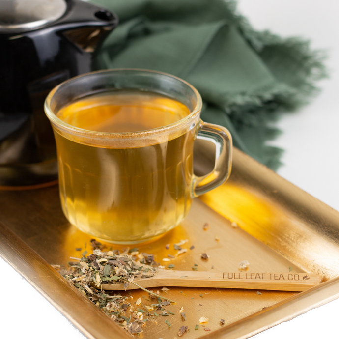 A glass cup of golden Organic Kidney Cleanse tea on a metallic tray beside a wooden spoon labeled ‘Full Leaf Tea Co.’ with scattered herbal tea leaves, and a dark teapot and green cloth in the background.