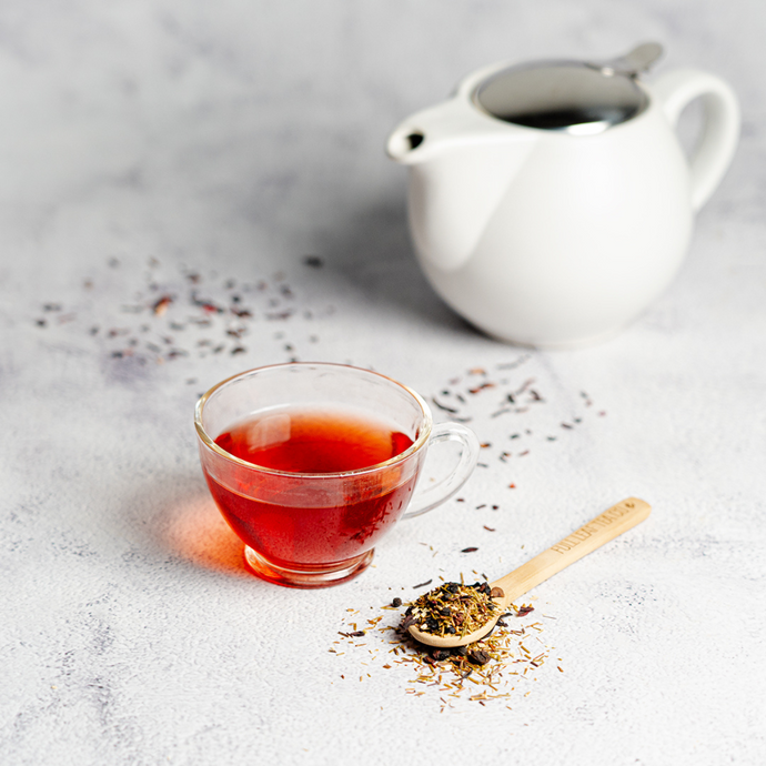 Cup of brewed Organic Vita Me Tea with a deep red hue beside a wooden spoon of loose herbal tea blend and a white teapot on a light stone surface.