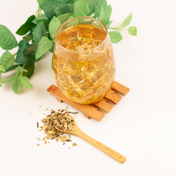 Glass of iced Organic Liver Cleanse Tea on a wooden coaster with loose leaf tea and a Full Leaf Tea Co. spoon beside fresh green leaves.