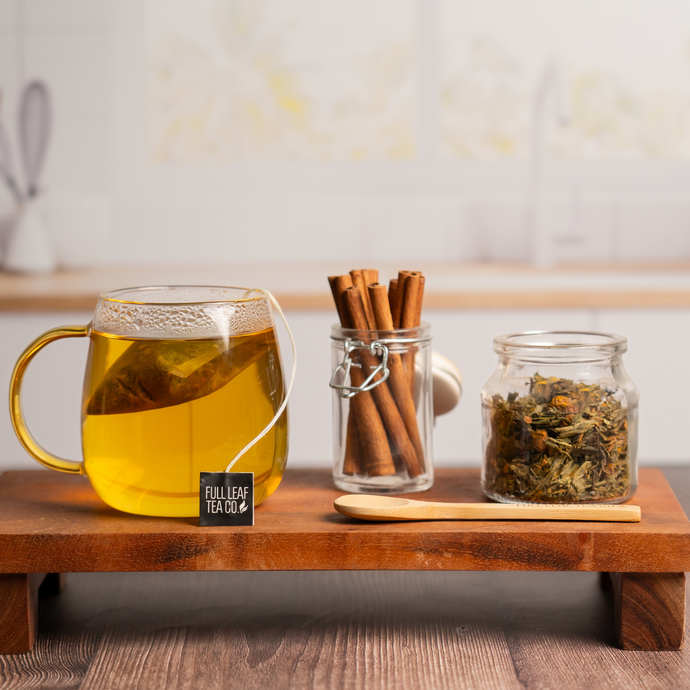 Glass mug of Organic Healthy Heart Tea steeping beside jars of cinnamon sticks and loose leaf tea on a wooden tray, with a wooden spoon and cozy kitchen background.
