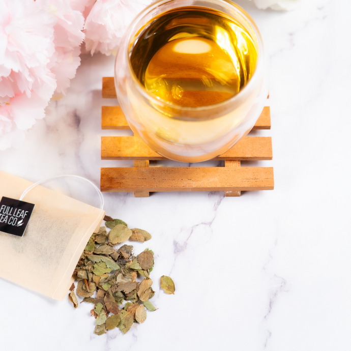 A cup of brewed Organic Bilberry Leaf tea on a wooden coaster beside a Full Leaf Tea Co. tea bag and dried bilberry leaves scattered on a white marble surface.