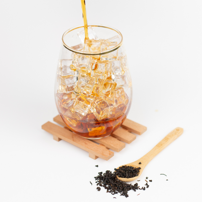Golden-brown Organic Assam iced tea being poured over ice cubes in a clear glass, with loose black tea leaves and a wooden spoon nearby.