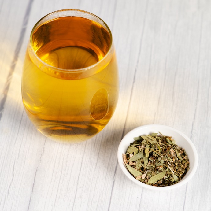 Glass of brewed Organic Healthy Colon Tea beside a small white bowl filled with loose herbal tea leaves on a light wood surface.