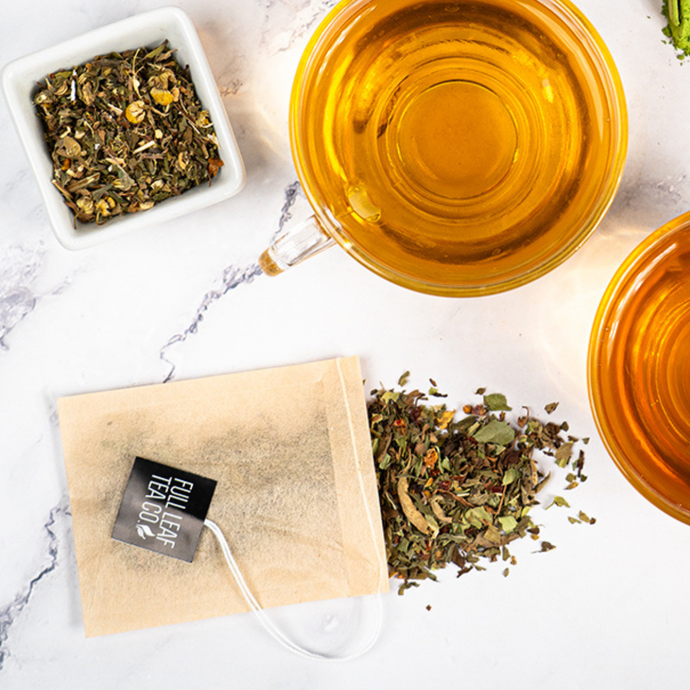 Top-down view of a tea setup featuring a Full Leaf Tea Company tea bag, loose leaf herbal blend, and two cups of golden-brown brewed tea on a marble surface.
