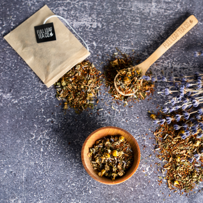 Loose leaf Organic Sleeping TranquiliTea blend with chamomile and lavender beside a wooden spoon, tea bag, and dried lavender flowers on a gray textured surface.