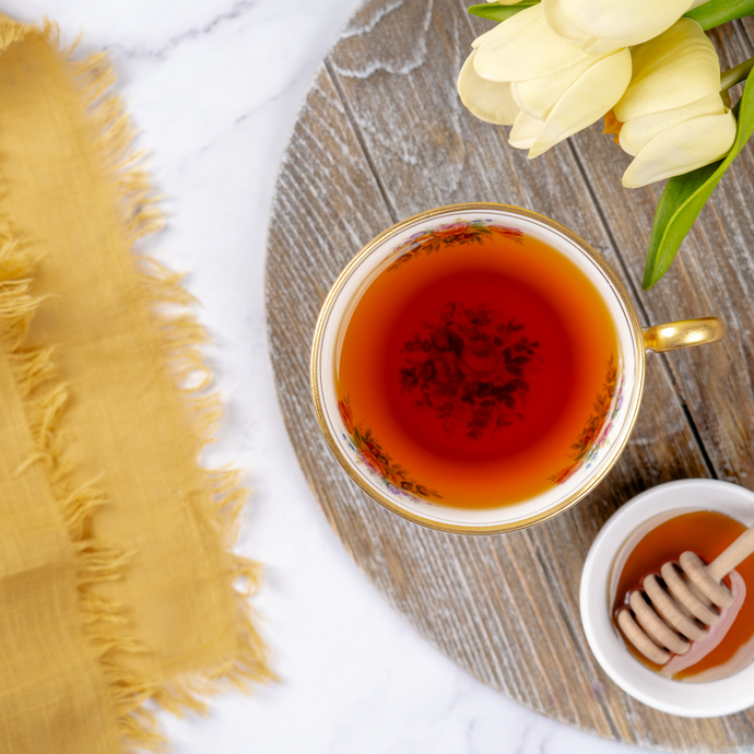 Cup of Organic Beauty Me Tea with rich amber color, served with honey dipper on a wooden tray, yellow tulips, and soft yellow cloth accent on marble background.