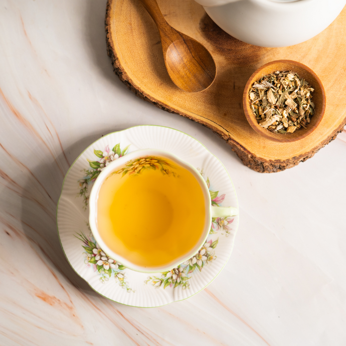 A floral teacup filled with golden herbal tea beside a wooden bowl of loose dried herbs and a spoon on a natural wood tray, creating a calm and soothing scene.
