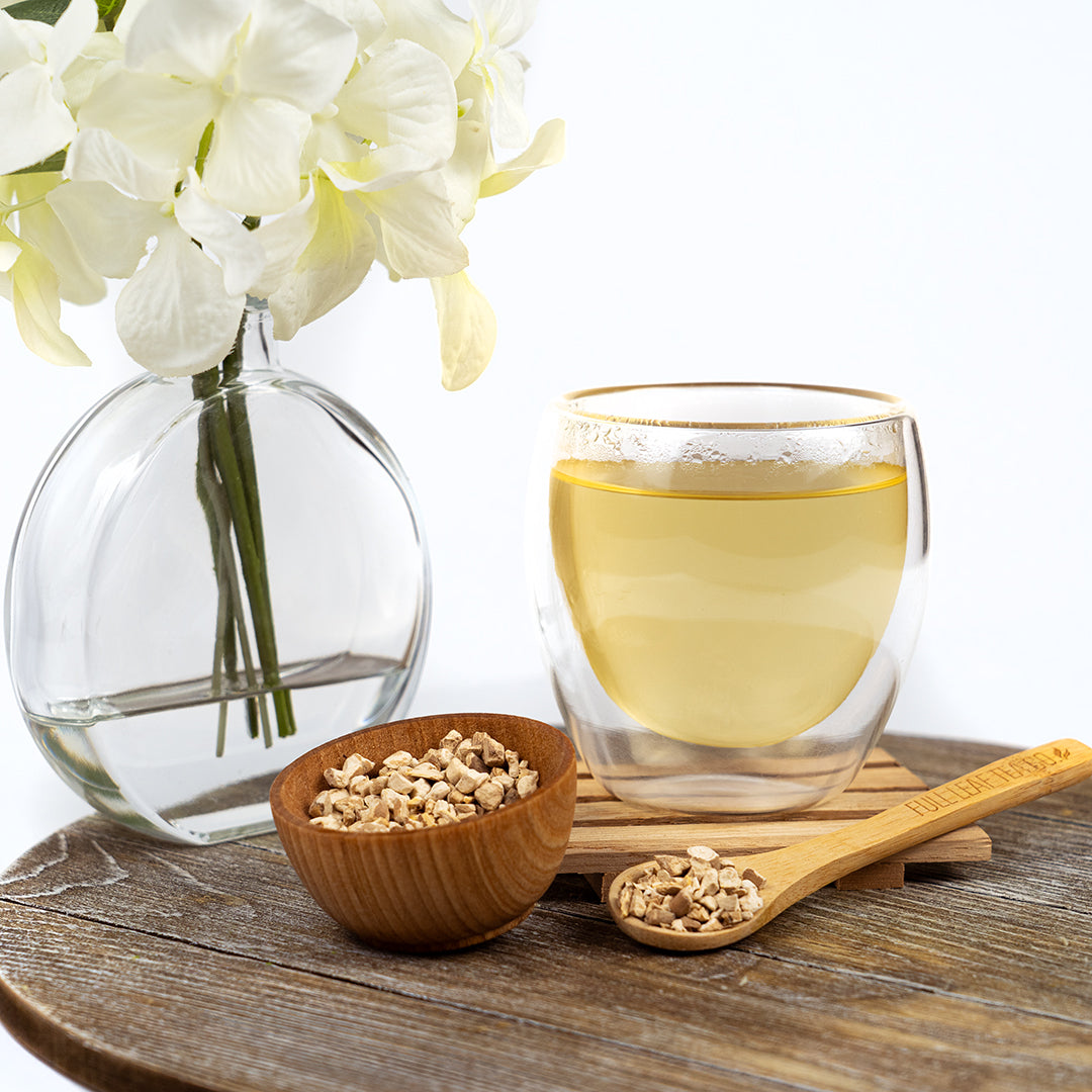 A clear mug filled with ashwagandha tea surrounded by white flowers, a brown cup filled with ashwagandha, and a Full Leaf Tea Co branded wooden spoon